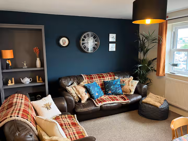 Cosy living room at Signal Box Cottage with deep petrol-blue walls, tartan wool throws over leather sofas, and warm brass lamplight.
