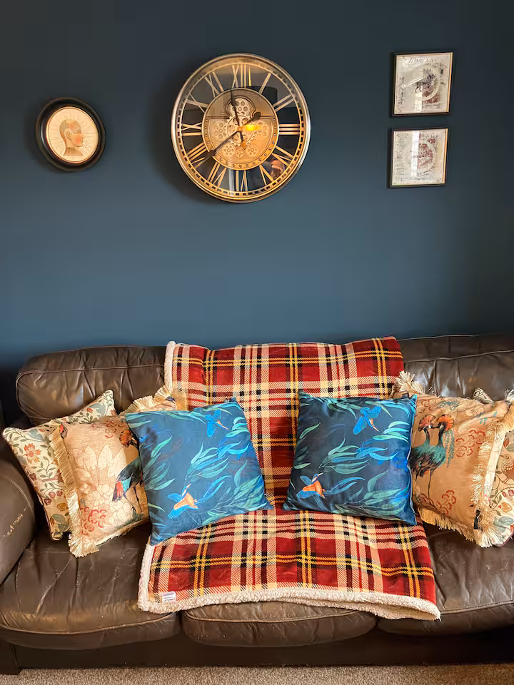 Close-up of the sofa styled with tartan throw, teal cushions and a brass carriage clock on the wall above.