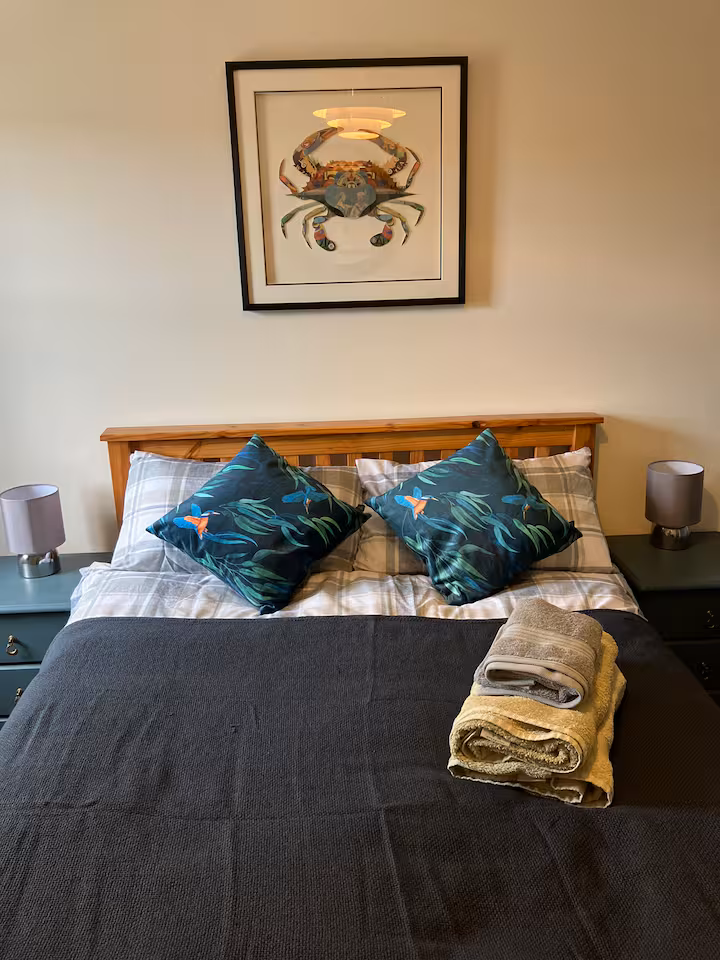 Bedroom with pine headboard, teal cushions, grey throw, and a framed crab print above the bed.