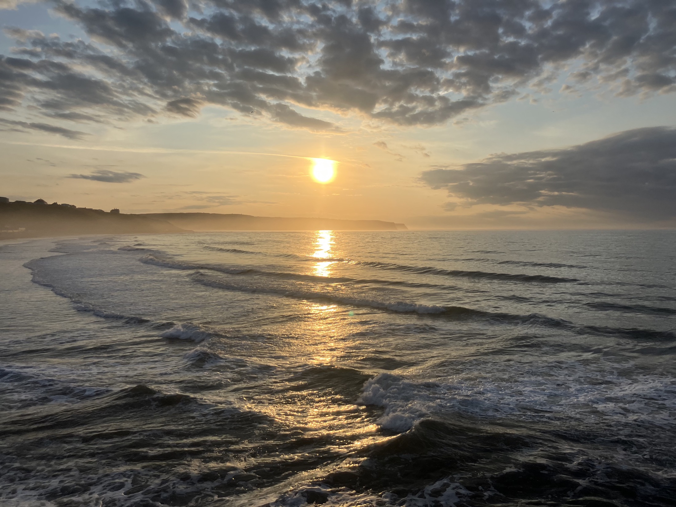 Golden sunset over the sea and waves along the Whitby coastline, with the headland silhouetted in the distance.